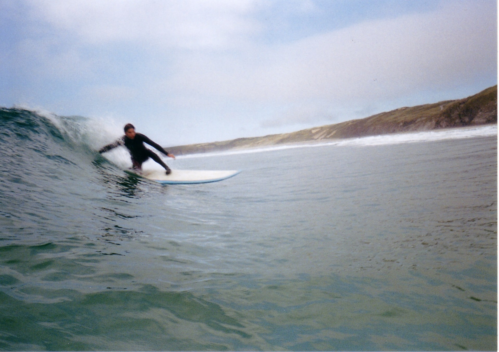 A surfer on a wave. He's crouching, touching the wave with his hand as the wave breaks. The wave is chest high, blue-green color. There are brown-green hills in the background on the land.